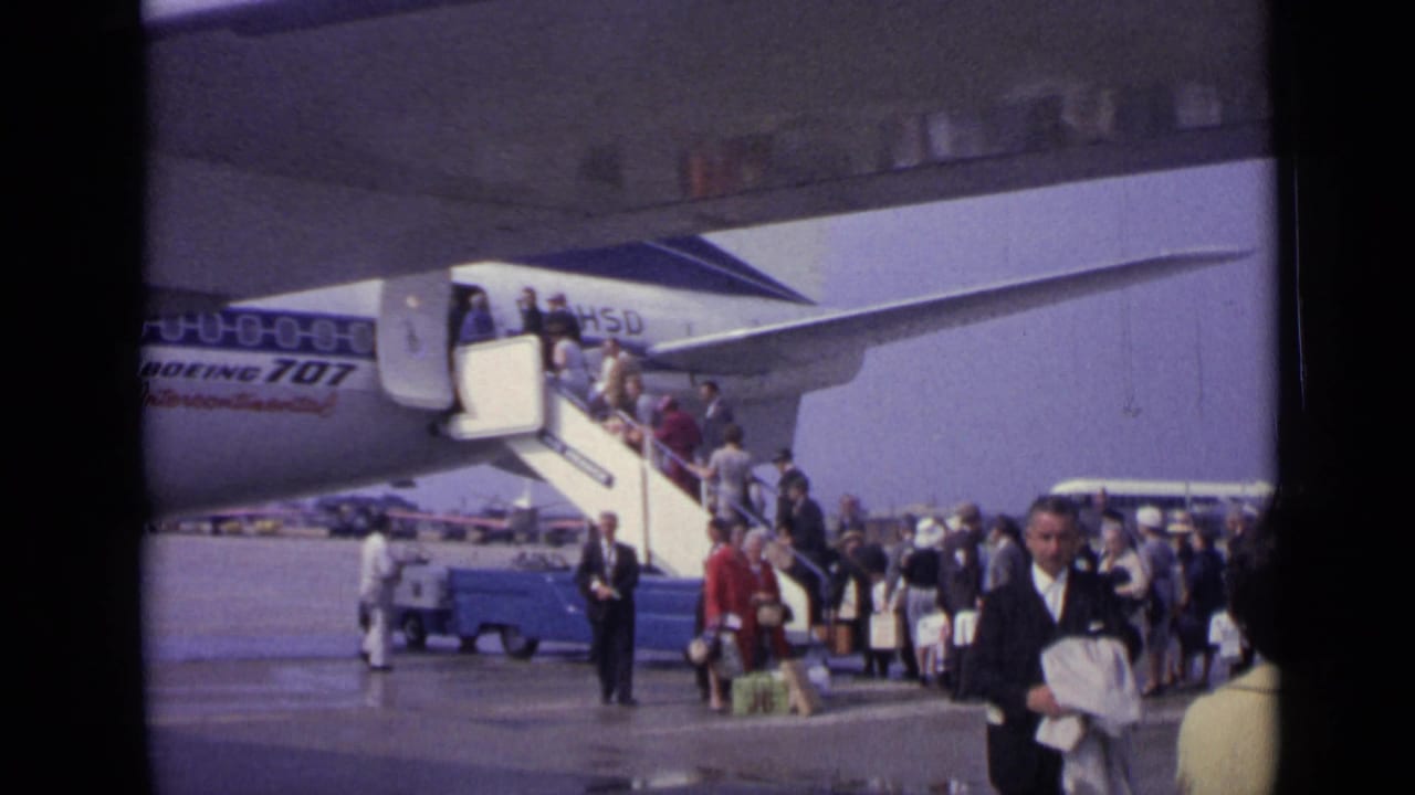 Passengers board a Boeing 707 at a London airport in 1962 while others wait below with bags and a small ground vehicle nearby.