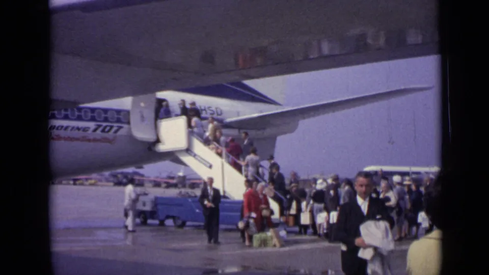 Passengers board a Boeing 707 at a London airport in 1962 while others wait below with bags and a small ground vehicle nearby.