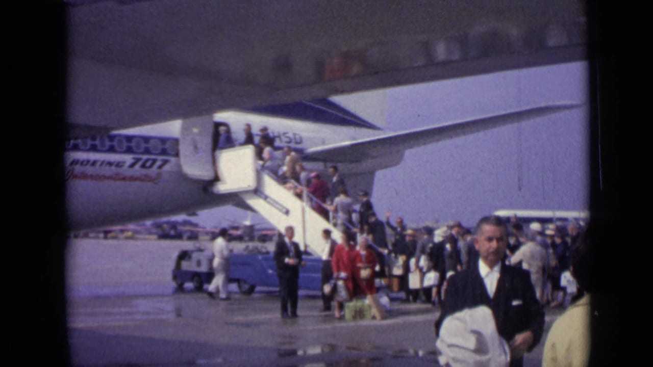 At a 1962 London airport, a suited man crosses the foreground while passengers board a Boeing 707 from mobile stairs behind him.