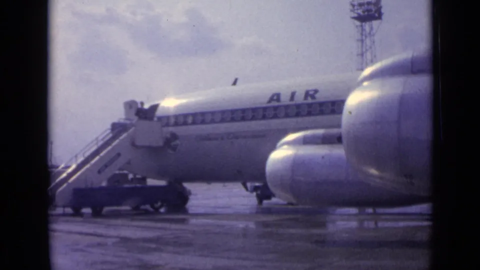 1962 London airport footage shows a passenger jet fuselage on wet tarmac, with mobile stairs at the door and partial blue lettering.