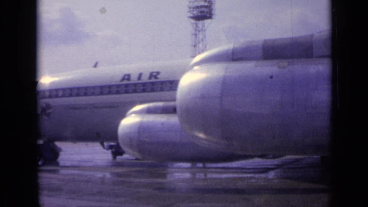A closer panned view shows two engine nacelles in the foreground, the passenger jet fuselage behind, and wet tarmac below.