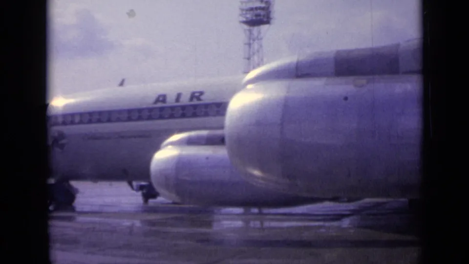A closer panned view shows two engine nacelles in the foreground, the passenger jet fuselage behind, and wet tarmac below.