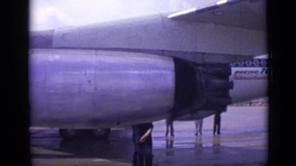 A Boeing 707 engine fills the frame at a London airport in 1962, with the aircraft fuselage and tarmac figures visible behind it.