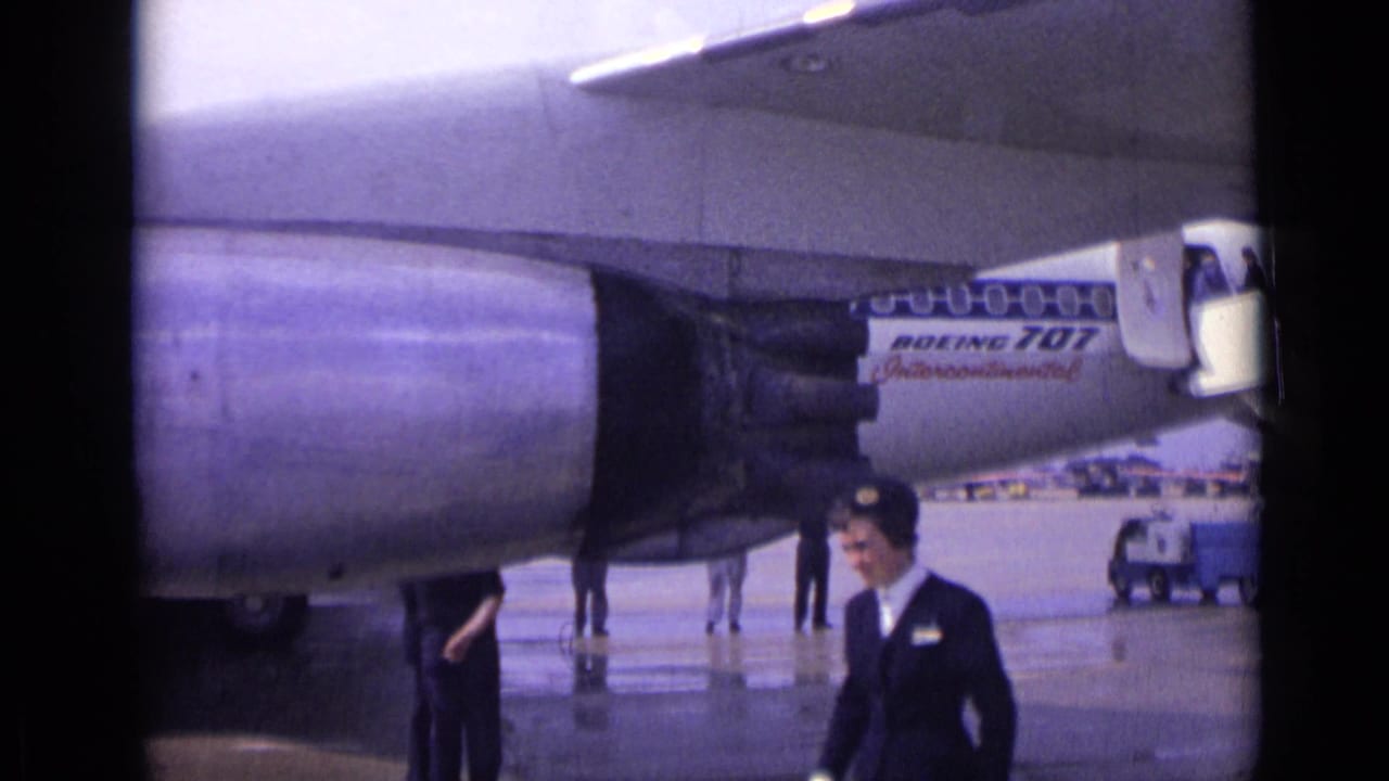 A uniformed flight attendant walks through the foreground near a parked Boeing 707 on wet London tarmac in 1962.
