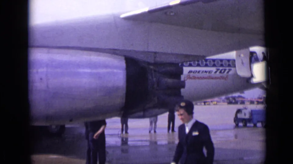 A uniformed flight attendant walks through the foreground near a parked Boeing 707 on wet London tarmac in 1962.