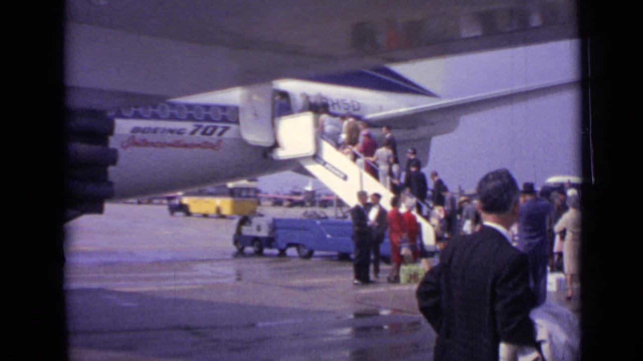 A home-movie view of passengers at mobile stairs beside a Boeing 707, with a suited man in the foreground on the tarmac.