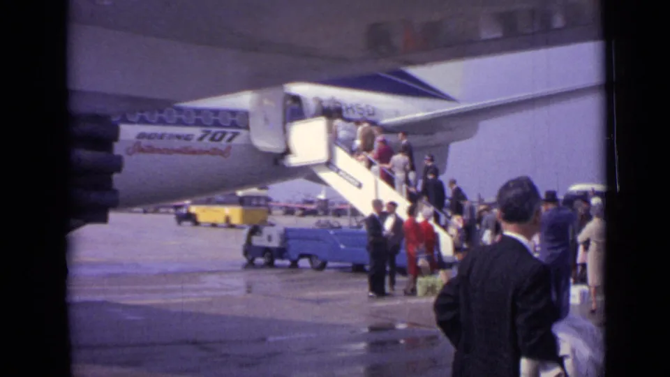 A home-movie view of passengers at mobile stairs beside a Boeing 707, with a suited man in the foreground on the tarmac.