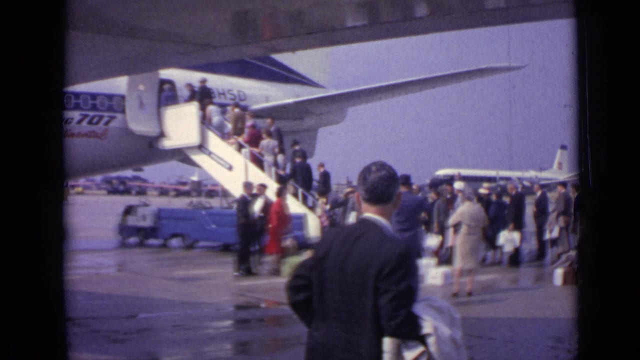 1962 London airport scene with passengers lined up at mobile stairs, service vehicles, wet pavement, and a distant aircraft.