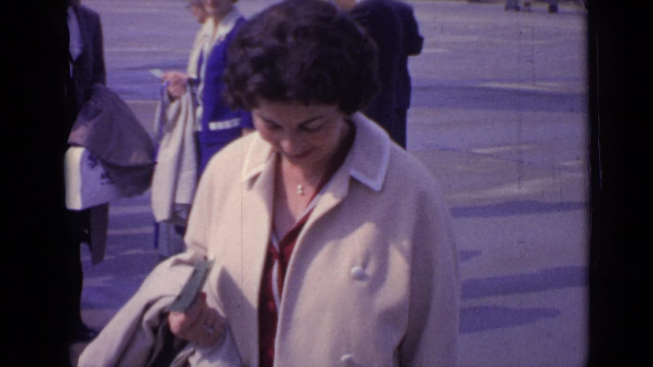 Woman in a cream coat looks down while holding an item on a paved street.