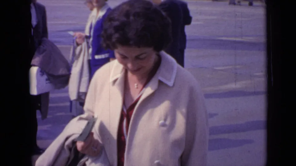 In 1962 London footage, a woman in a cream coat and red dress looks down while standing among pedestrians.
