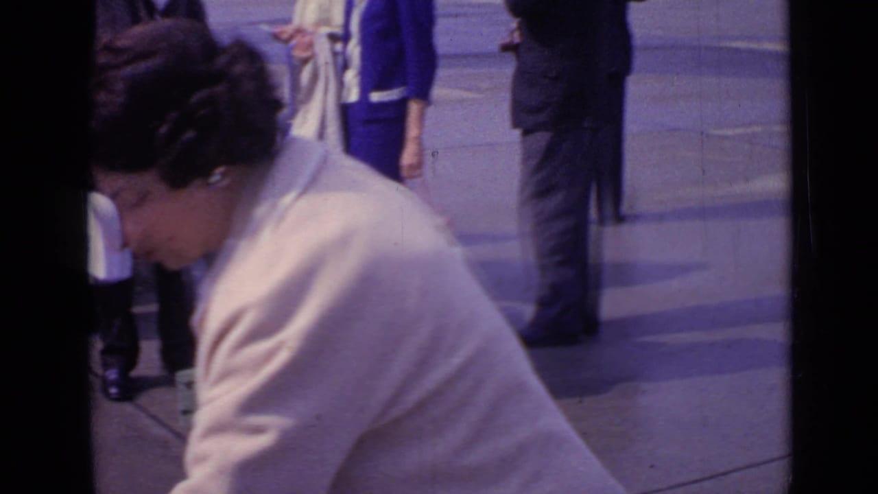 The woman bends forward in a cream coat as other pedestrians stand or walk behind her in the London street scene.