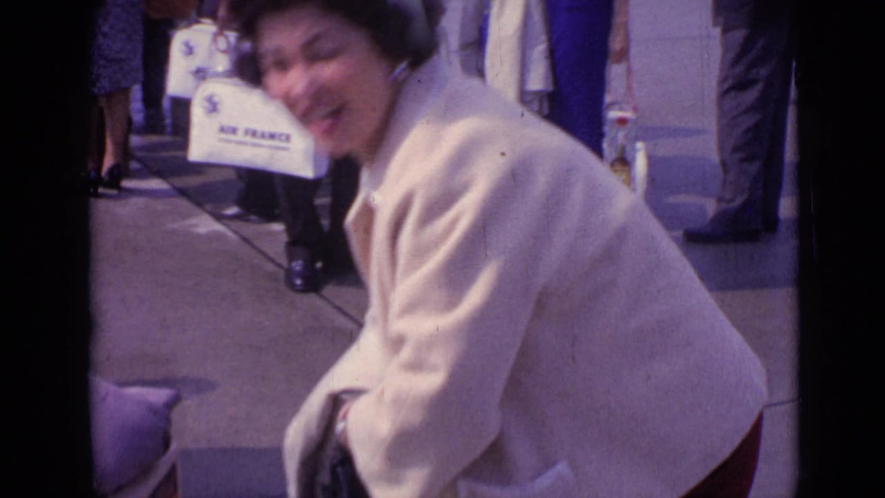 A woman in a cream coat bends near the camera, with white Air France luggage and pedestrians visible in the background.