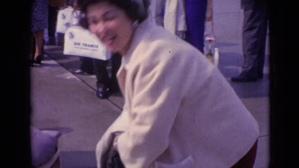 A woman in a cream coat bends near the camera, with white Air France luggage and pedestrians visible in the background.
