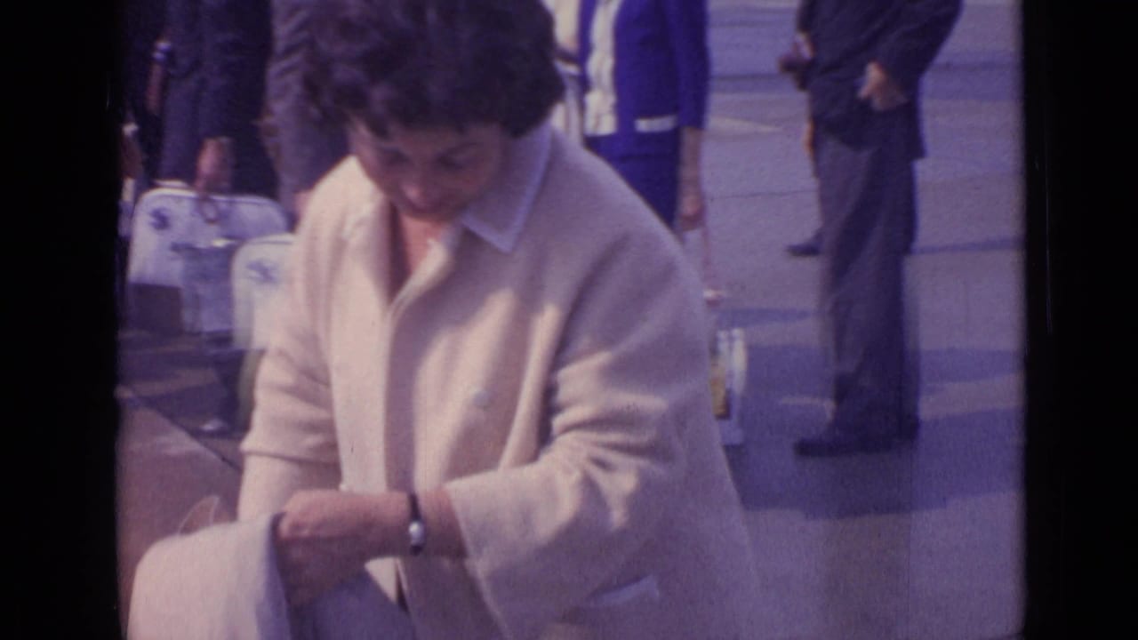 The woman looks down while handling a bag or item, with pedestrians and white luggage behind her in 1962 London footage.