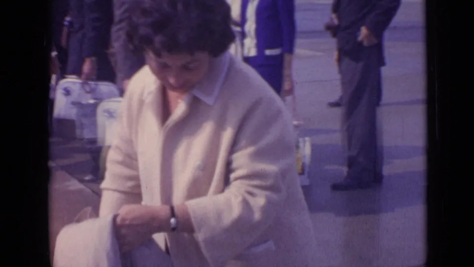 The woman looks down while handling a bag or item, with pedestrians and white luggage behind her in 1962 London footage.