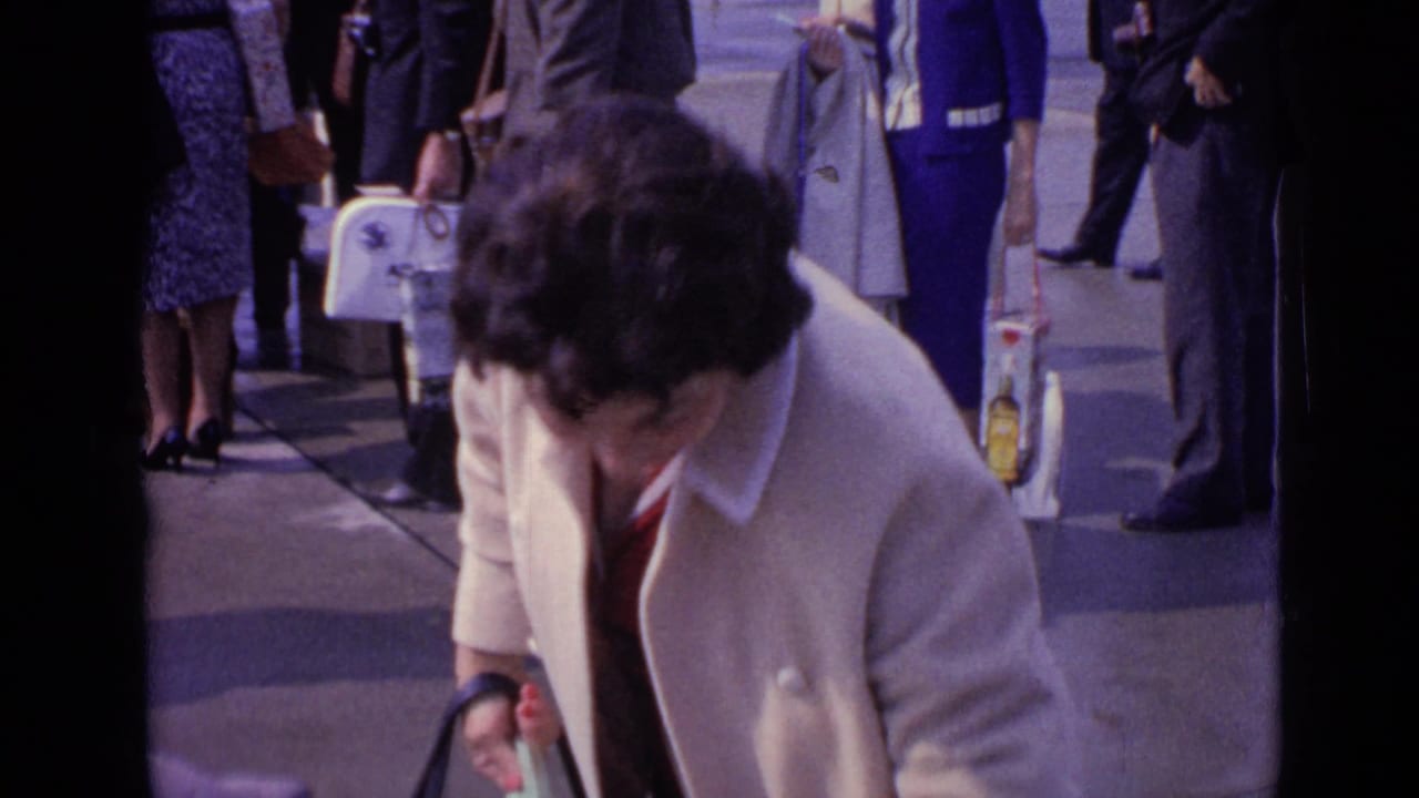 Woman in a cream coat bends slightly among travelers on a sidewalk.