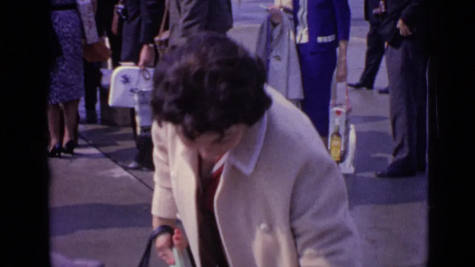 In a 1962 London home-movie frame, a woman in a cream coat stands near travelers with handbags and luggage on a busy pavement.