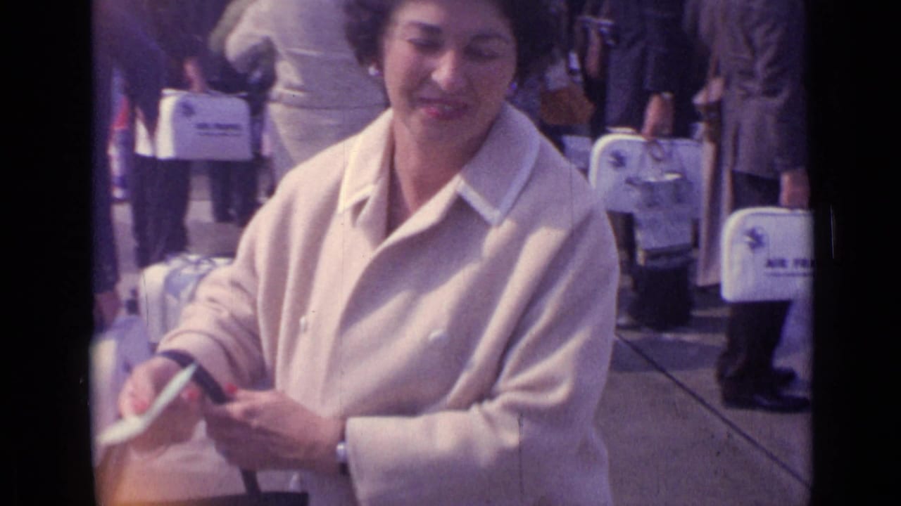 A smiling woman in a cream coat adjusts a small bag while standing among other travelers in the 1962 London home-movie scene.