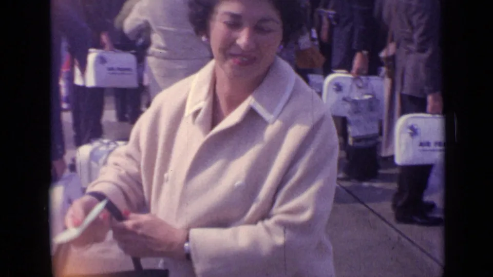 A smiling woman in a cream coat adjusts a small bag while standing among other travelers in the 1962 London home-movie scene.
