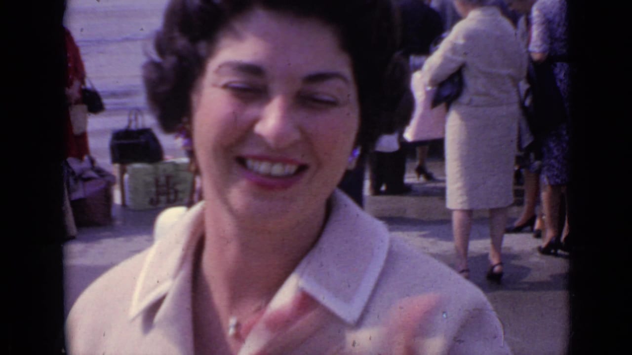 Close handheld view of the smiling woman in a cream coat, with travelers and luggage visible behind her in London according to clip metadata.