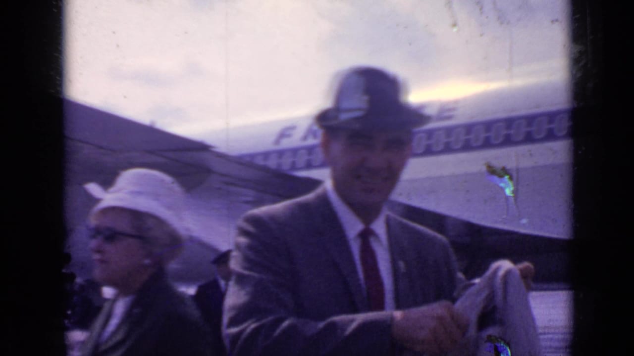 A man in a suit and fedora stands near an aircraft fuselage with partial lettering visible, with another traveler in a hat nearby.