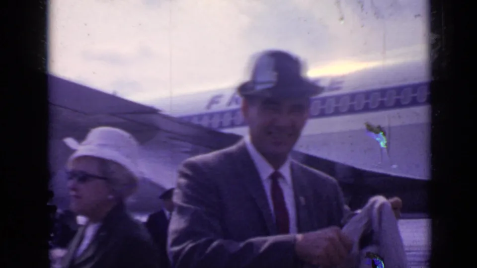 A man in a suit and fedora stands near an aircraft fuselage with partial lettering visible, with another traveler in a hat nearby.