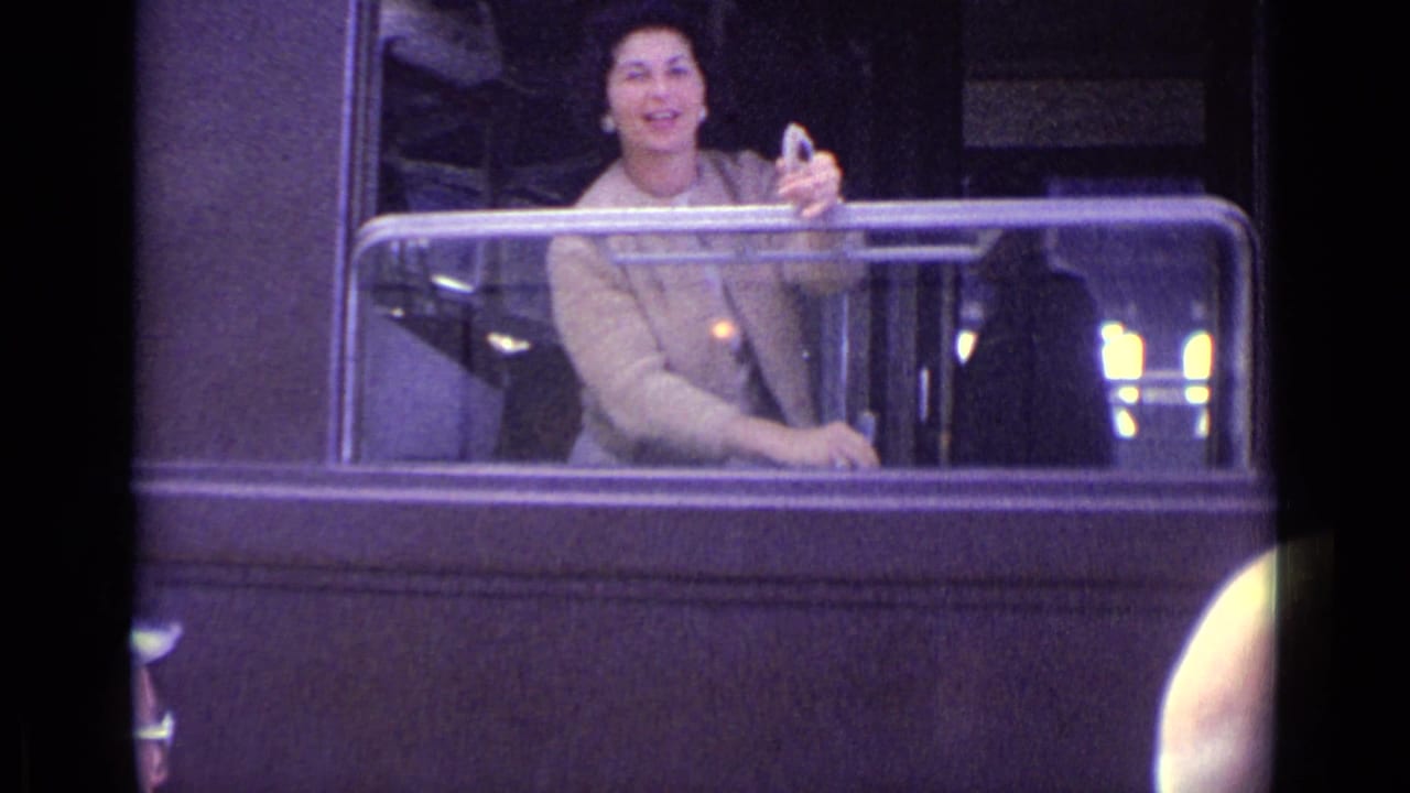 A smiling woman in a brown coat leans from the train window, holding a glass during a 1962 Lucerne travel moment.