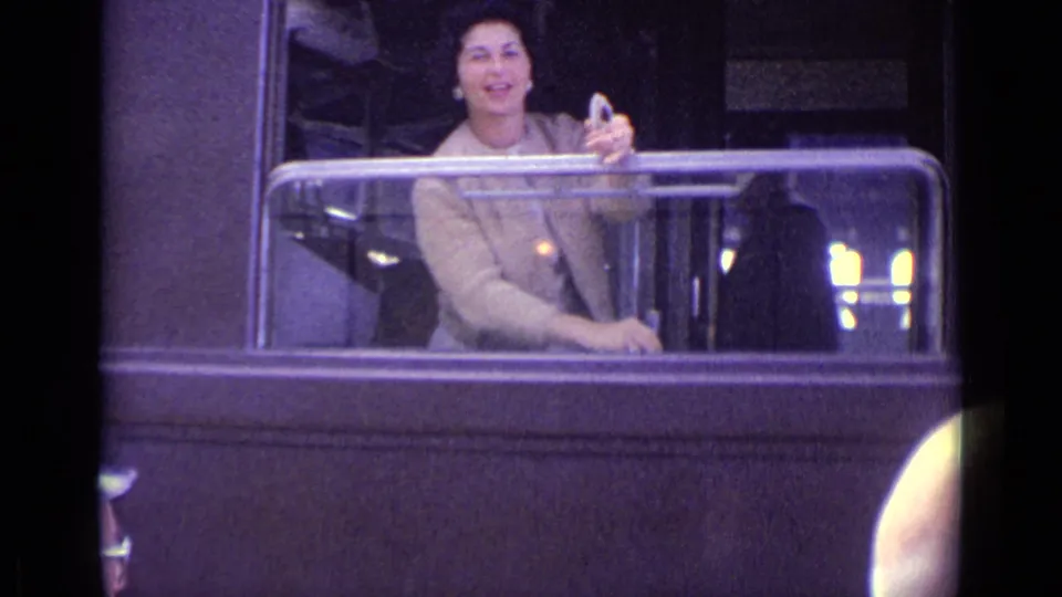 A smiling woman in a brown coat leans from the train window, holding a glass during a 1962 Lucerne travel moment.