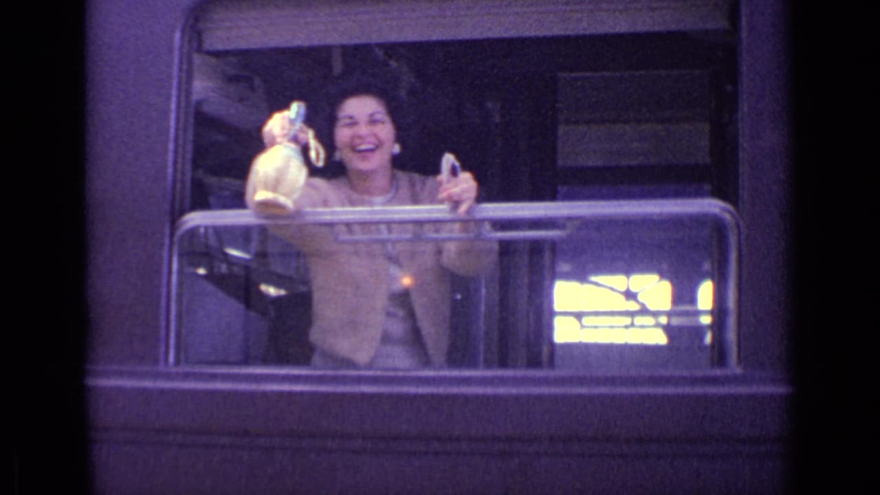 The woman smiles from the train window and extends a bottle while holding a glass in the Lucerne 1962 home movie clip.