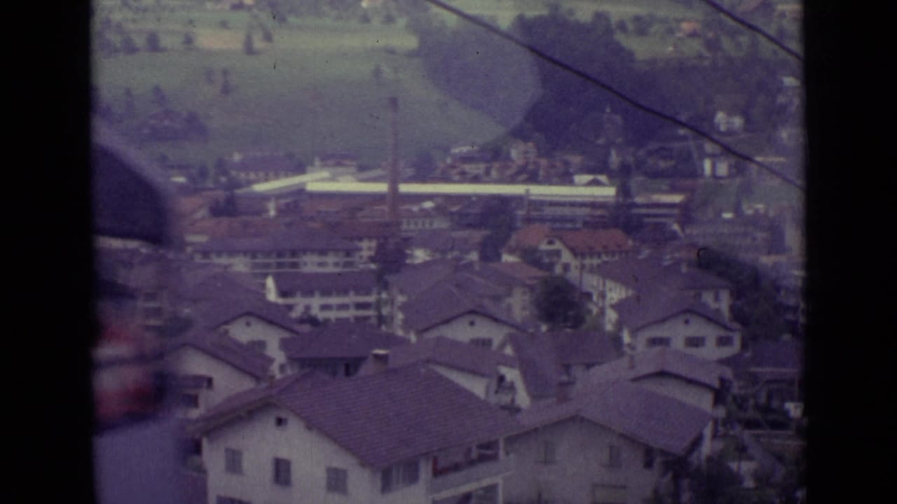 Elevated view over rooftops, hillside fields, overhead cable, and distant buildings.