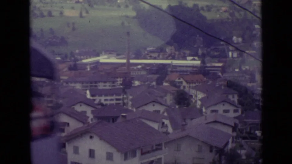 Elevated 1962 Lucerne view across residential rooftops, hillside fields, trees, overhead cable lines, and distant buildings.