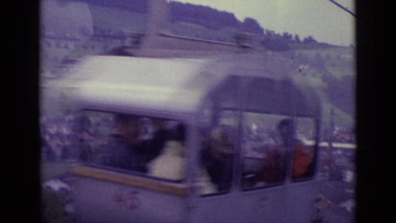 A silver cable car with visible passengers moves past the camera in 1962 Lucerne, with hillside rooftops and cables behind it.