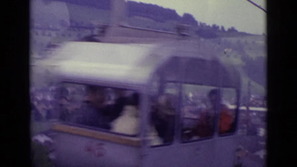 A silver cable car with visible passengers moves past the camera in 1962 Lucerne, with hillside rooftops and cables behind it.