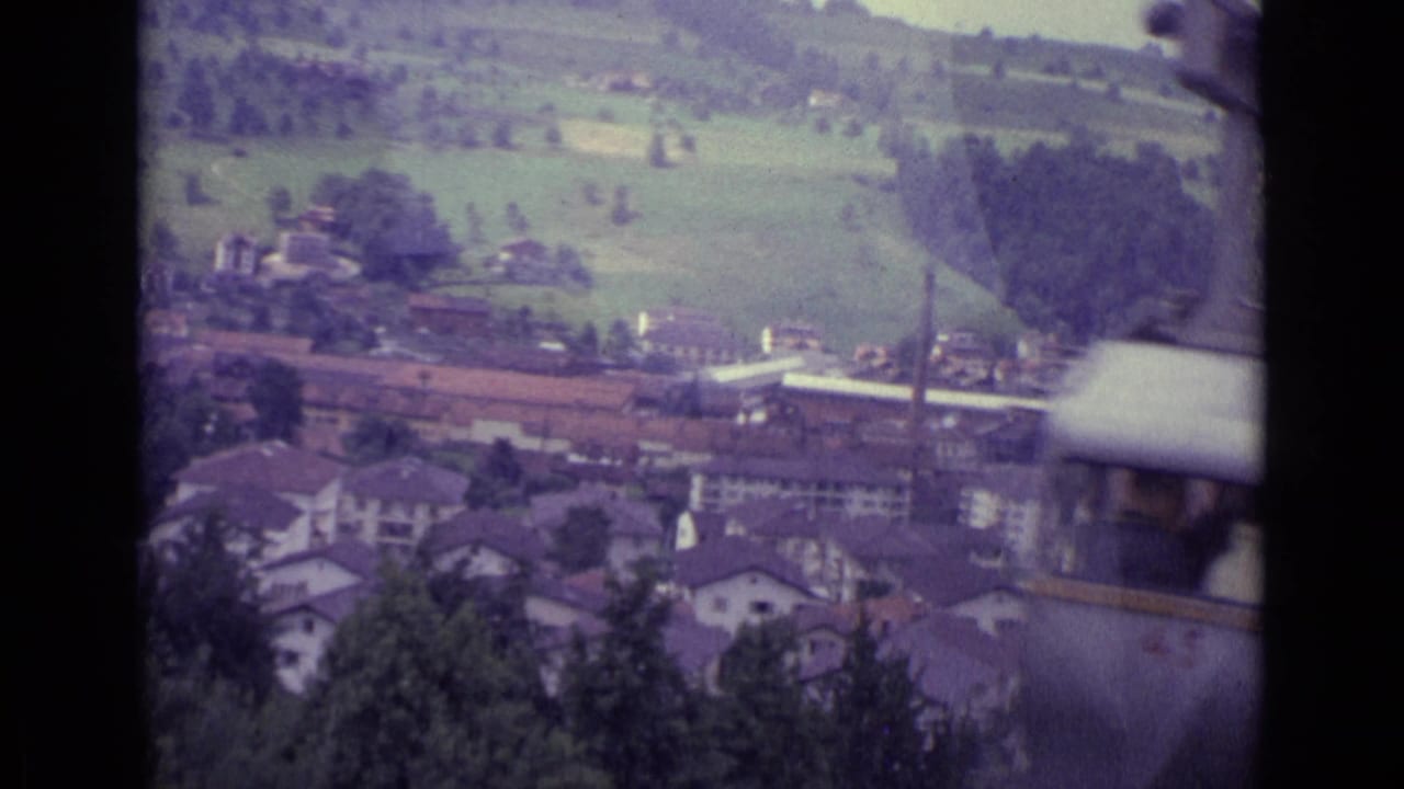 Cable-car view over hillside houses, fields, trees, and a valley town seen through dark window bars.