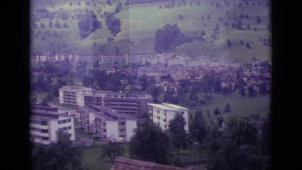Elevated 1962 Lucerne view with apartment blocks, trees, hillside fields, and village buildings softened by 8mm film color.