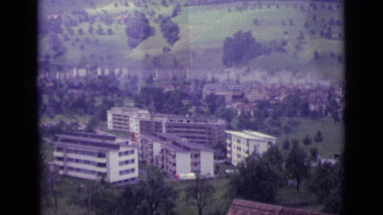 Swiss hillside town with apartment buildings, trees, and green slopes in a faded 8mm frame.