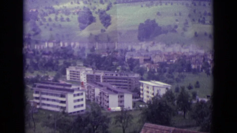 1962 Lucerne home-movie view of apartment blocks below lush hills, with trees and a hazy townscape in the distance.