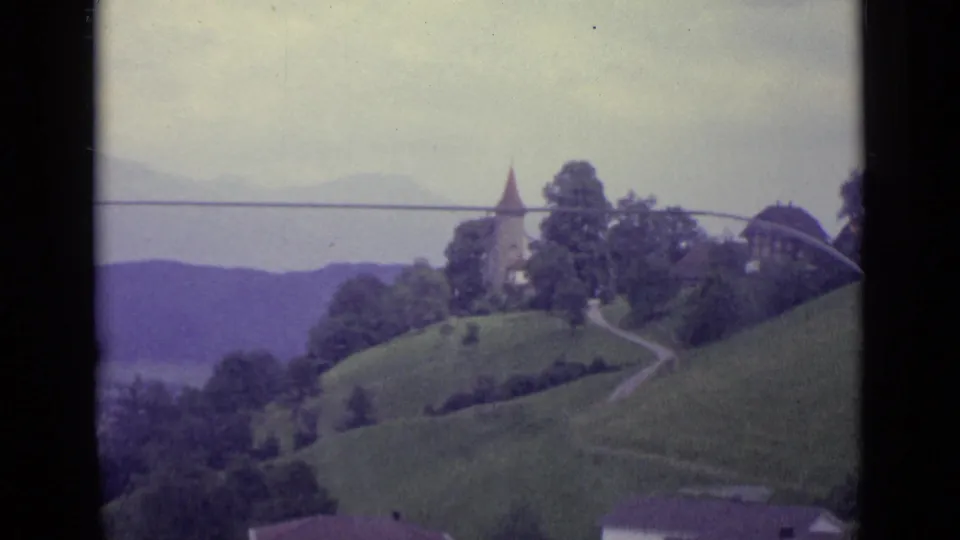 1962 Lucerne home-movie frame showing a church tower on a grassy hillside with trees and a path against a hazy backdrop.