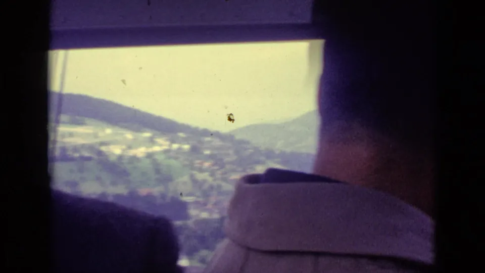 Inside a Lucerne-area cable car in 1962, two adult men face the windows as hills and a village appear outside.