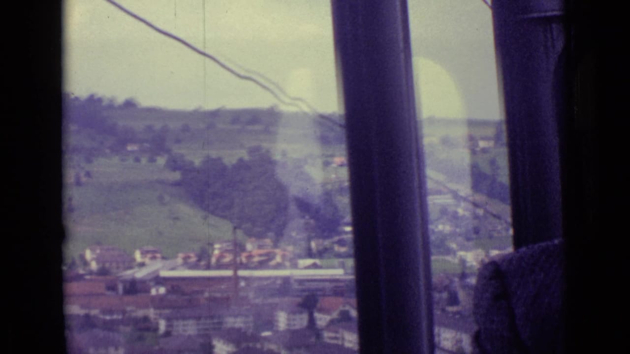 1962 Lucerne-area hillside, buildings, fields, and a smokestack viewed through dark cable-car window bars.