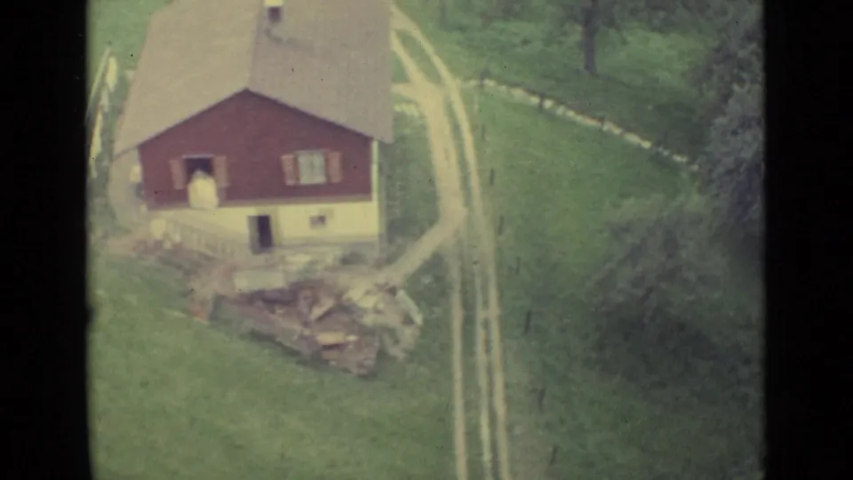 1962 Lucerne-area rural house with red upper walls, pale lower level, grassy slope, and winding road below the cable car.