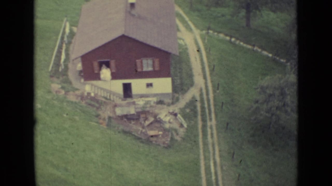 Red country house on a grassy hillside with a dirt track and a person near the doorway.