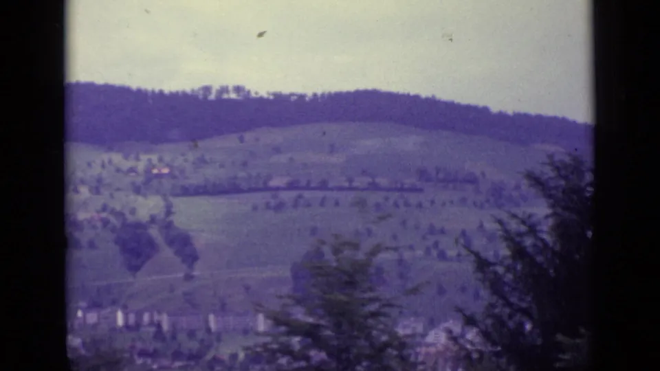 Lucerne-area hillside view from the 1962 8mm clip, with scattered trees, fields, and small distant buildings.