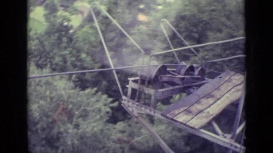 1962 8mm frame showing cable-car wheels, wires, and a steep support structure above wooded green terrain near Lucerne.