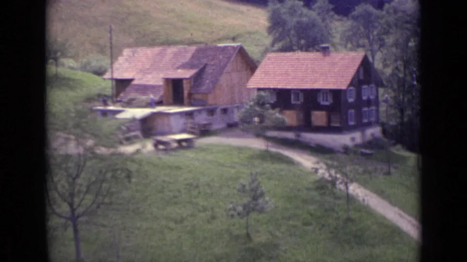 1962 Lucerne frame of two wooden houses with red roofs, seen across a green valley with scattered trees and 8mm frame edges.
