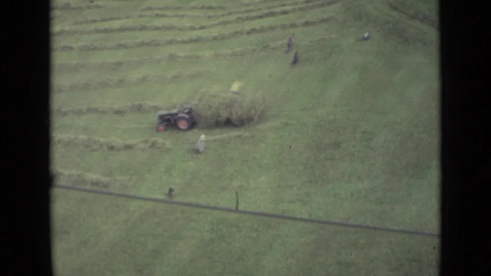 Aerial home-movie view in Lucerne, Switzerland, showing a small tractor beside cut hay and workers moving across the field.