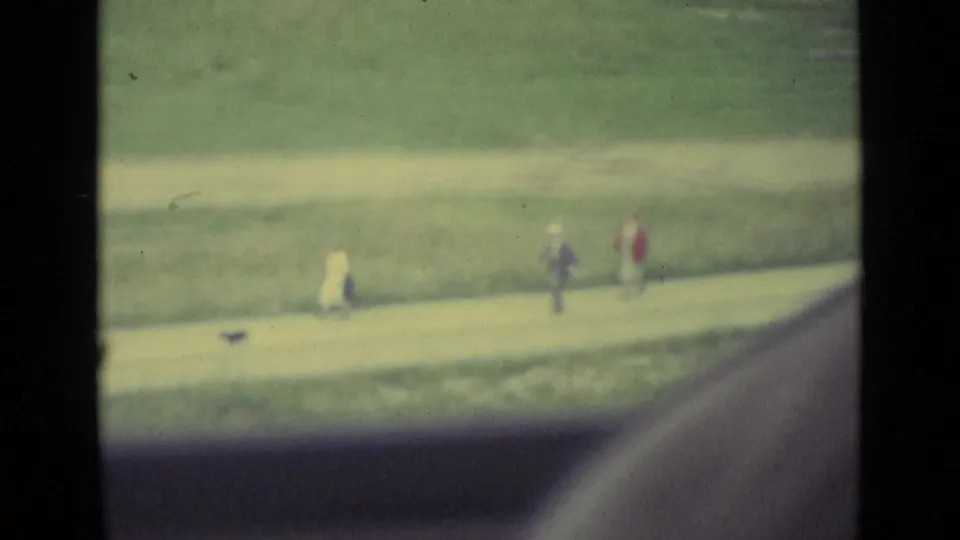 Lucerne 1962 home-movie frame with several distant pedestrians on a path beneath a grassy hillside.