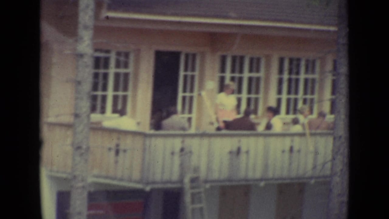 People sit and stand on a balcony of a white building with tall windows.