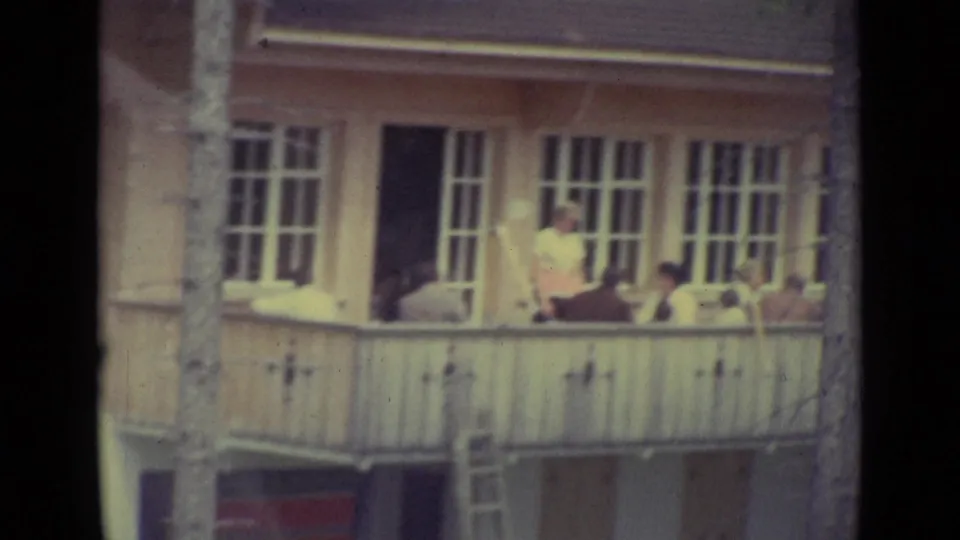 1962 Lucerne home-movie frame showing people gathered on an upper balcony of a white building with tall windows.