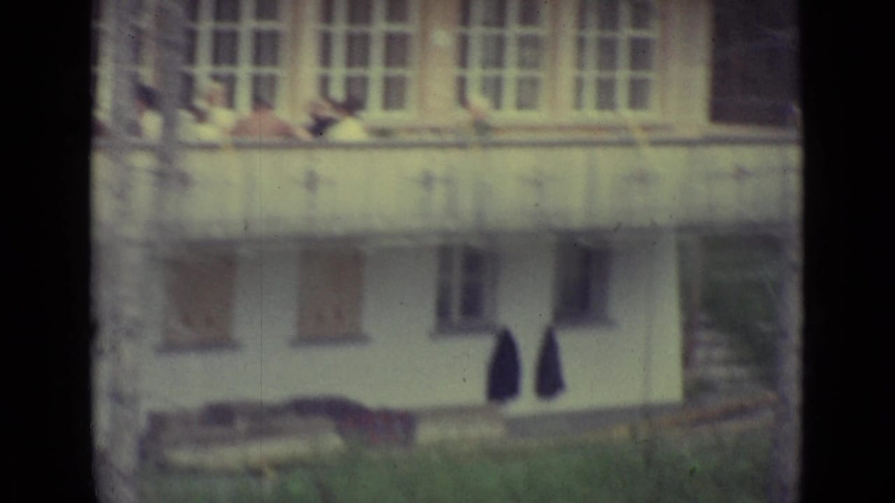White building exterior with balcony above, shuttered windows below, and garments hanging outside.